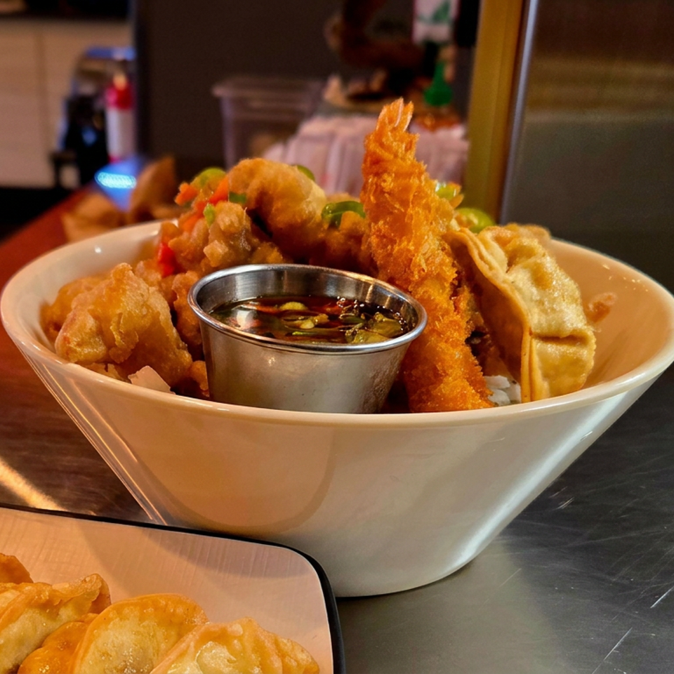 Bowl of crispy Asian appetizers with shrimp, dumplings, vegetables, and dipping sauce on a restaurant counter.