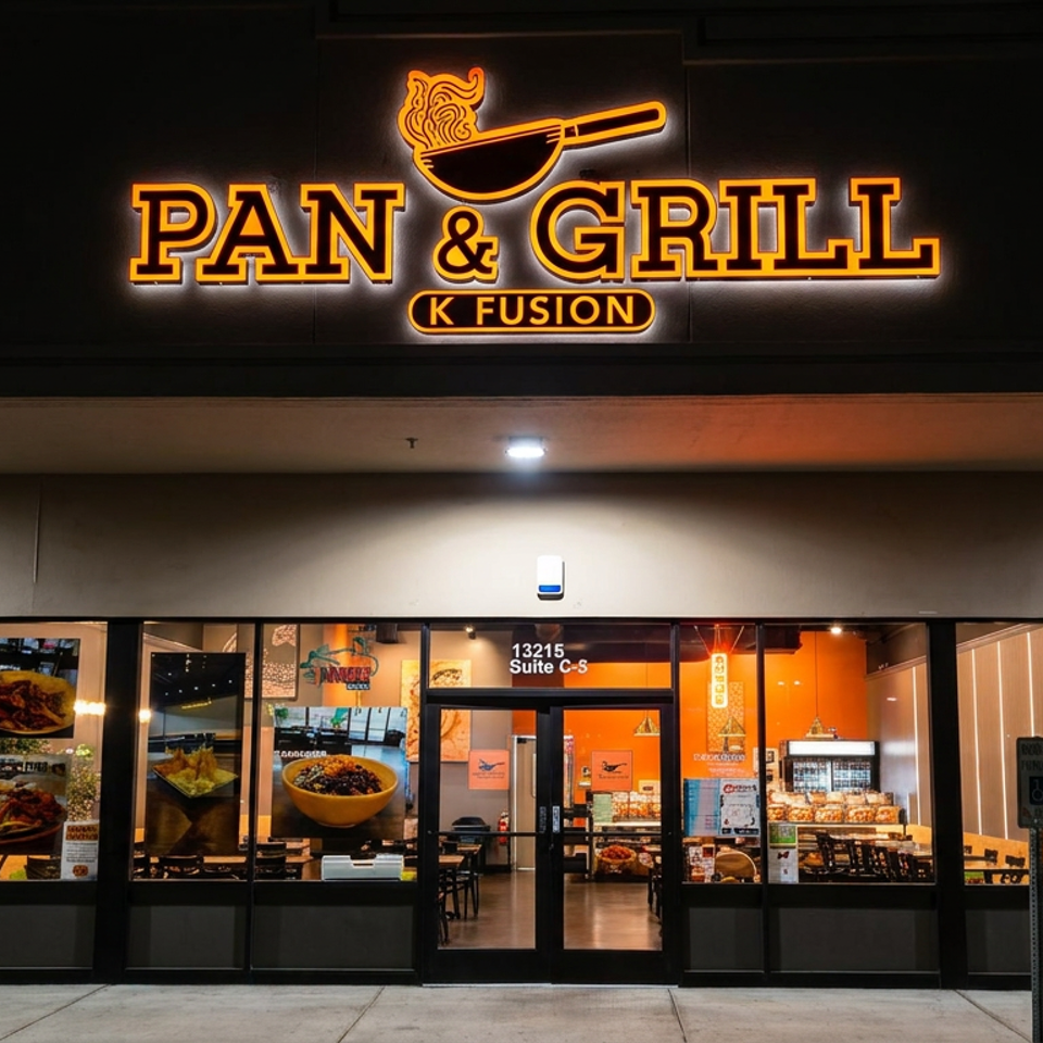 Exterior of Pan & Grill K Fusion restaurant with glowing sign, glass storefront, food displays, and warmly lit dining area.